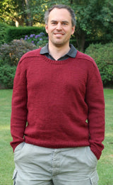 Man stands in a green backyard. He wears a cranberry red sweater over a charcoal button up shirt and khaki pants.  thumbnail