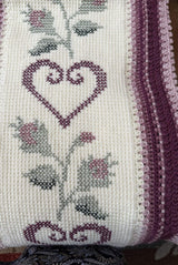 Close up of afghan. The background is cream, with a lighter and darker pink border. The cream panel has dark pink hearts and flower buds and green stems cross stitched on.  thumbnail