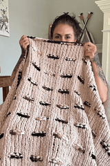 Woman holds a knit afghan up over her chin. The blanket is a warm tan with dashes of darker brown.  thumbnail