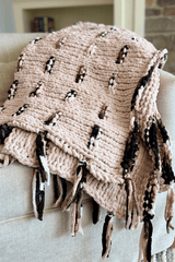 Knit afghan draped over beige armchair. The blanket is knit in tan chenille yarn and has darker brown multicolor dashed stripes and tassels on edge.  thumbnail