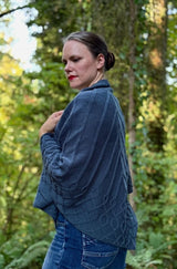 Lower side view of woman wearing draped shawl and jeans in a green forest. The shawl has wider vertical ribs and a cabled lower border.  thumbnail