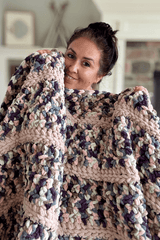 Woman holds crochet blanket up to chin. The blanket is made in stripes of multi-color yarn and a pale beige.  thumbnail