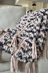 Crochet blanket draped over armchair, showing wide stripes of multi-color chenille yarn and thin stripes of tan yarn, with tassels.  thumbnail