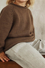 Cropped side view of warm brown knit sweater on a toddler, wearing striped pants and sitting on a bench.  thumbnail