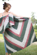 Woman standing in a green yard holds a crochet afghan. The blanket is made in whisper green, medium green, and a coral pink.  thumbnail