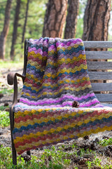Crochet afghan draped over wooden bench in a wooded area.  thumbnail