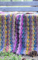 Close up of crochet blanket laid across seat of wooden bench. The blanket has a scallop shell pattern worked in purple and yellow yarns, with pops of blue and rusty red.  thumbnail