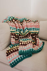 Crochet blanket draped over a beige couch. The blanket has textured stripes in kelly green, aqua, pink, butter yellow, cream, and dark brown.  thumbnail