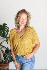 Front view of woman wearing yellow crochet top and jeans, standing in front of a cream wall with a potted plant behind her.  thumbnail