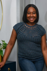 Front view of woman wearing a slate blue crochet top with jeans. She stands in the corner of a room with one hand resting on a chest of drawers, with a potted plant and mirror.  thumbnail