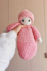 Crochet baby doll held up by hand of person off camera. The doll has a round head, arms, and a floppy sack body. The body and hat is pint and the face and hands are cream.  thumbnail