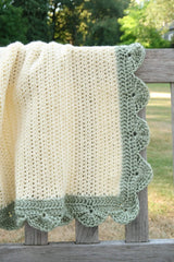 Crochet blanket draped over back of wooden bench. The blanket body if cream and it has a sage green scalloped edge.  thumbnail