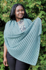 Woman wears a knit rectangular shawl over a top and dark pants, standing in front of a green tree.  thumbnail