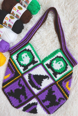 Flat lay of crochet bag on cream rug with yarn balls in the top left. The bag is made of squares with black cats, witch hats, ghosts and a spellbook.  thumbnail