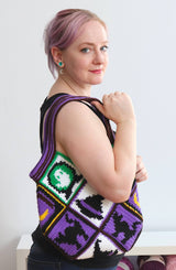 Woman wears crochet bag over her shoulder  - the bottom of the bag reaches her hip. The bag is made in purple, white, and green with yellow accents.  thumbnail