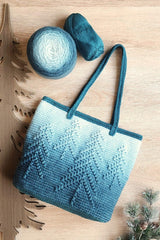 Crochet tote bag on pale wooden surface, with a pine tree cutout and pine bough in the background. The bag fades from dark teal at the base to near-white at the top, with a darker top edge and straps. The bag has pine tree relief design.  thumbnail