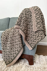 Crochet afghan draped over the arm of a couch. The afghan is olive green and warm tan with an all over mosaic design.  thumbnail