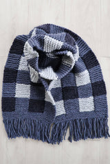 Knit scarf laid on pale wood background. It's knit in a gingham pattern in three shades of blue, with fringe on the ends.  thumbnail