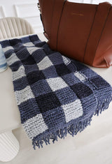 Gingham knit scarf laid on counter with a brown shoulder bag in the background.  thumbnail