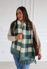 Woman wearing crochet scarf over sweater and jeans. The scarf ifs made in a gingham design in two shades of green plus cream, with fringe on the ends. It hangs to low hip when draped around the neck.  thumbnail