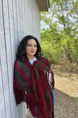 Woman leans against side of wooden structure, with trees in the background. She is wrapped in a large knit shawl with an all over lace pattern and long fringe.  thumbnail