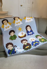 Afghan craped over grey couch indoors. The afghan has a pale blue background with three rows of cartoon style figures from the nativity scene.  thumbnail