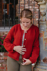 Woman wearing a red-orange cardigan leans against a pillar with a brick wall and iron gate in the background.  thumbnail