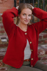 Woman sitting on brick steps wearing red-orange cardigan. She has her arms raised and the cardigan its buttoned at the very top.  thumbnail