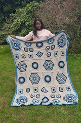 Woman standing in green backyard holding crochet afghan. The blanket is made of squares of many sizes, with a cream background and varied blue motifs.  thumbnail