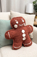 Crochet gingerbread man pillow sitting on a cream armchair. The pillow is warm brown with white accents.  thumbnail