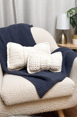 Two crochet pillows in the shape of bows sit on a blue blanket draped over a cream armchair.  thumbnail