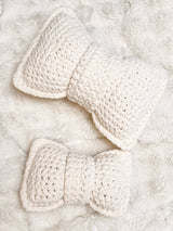Top down view of two crochet bow pillows, one small and one large, in cream, laid on a fuzzy cream blanket.  thumbnail