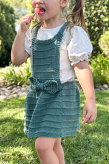 Cropped front view of young girl wearing knit pinafore in a dark sage green over a white t-shirt. The pinafore has a knee-length skirt and a bow at the waist.  thumbnail