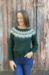 Woman in a green sweater with cream yoke design and blue jeans, standing in front of a weathered wooden barn door.  thumbnail