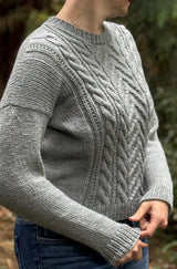 Side view of woman wearing grey sweater with cables on the front and plain sleeves, sides, and back. The sweater has long sleeves and a drop shoulder construction.  thumbnail