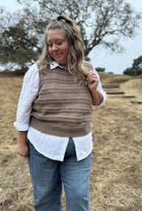 Woman standing on a hill with golden grass. She wears a knit vest with horizontal bands of texture over a white shirt and jeans.  thumbnail