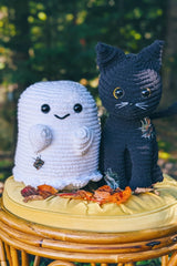 Crocheted ghost and black cat sit on a yellow stool in front of trees. They both have sewn-on patches, plastic eyes, and embroidered features.  thumbnail