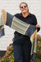Woman in black top and dark jeans stands in front of a white painted wall and hydrangea bush. She holds up a knit shawl or scarf that is color blocked in denim blue, sage green, and golden yellow, with each block separated by cream.  thumbnail
