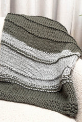 Knit blanket draped over cream armchair, in front of a cream drapery. The blanket is color-blocked in grey-green and pale grey.  thumbnail