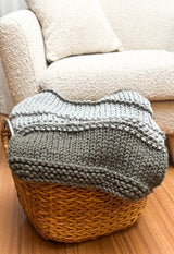 Knit blanket folded in wicker basket, sitting in front of cream armchair.  thumbnail