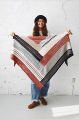 Woman in a black hat, white sweater, and jeans standing in an all-white room. She holds a knit shawl toward the camera. The shawl is made of blocks of different textures in terracotta, cream, pale blue, beige, and black.  thumbnail