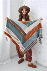 Woman standing in an all-white room holding a shawl toward the camera. She wears a flat brimmed hat, cream shirt, and rust red pants. The crochet shawl is color blocked in rusty orange,  dark sage green, cream, chocolate brown, and pale blue.  thumbnail