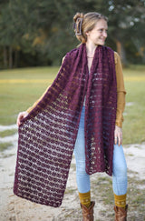 Woman standing in an open grassy area with trees behind. She wears a large lacy crochet stole in a dark red-purple color, over a tan top and light colored jeans.  thumbnail