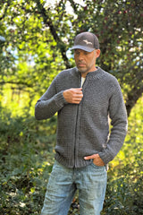 Man standing in a wooded area, wearing a zippered crochet cardigan, baseball hat, and jeans.  thumbnail