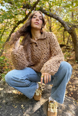 Woman crouched on boulder in early fall woods. She wears sneakers, jeans, and a crochet pullover with long sleeves and a polo collar.  thumbnail