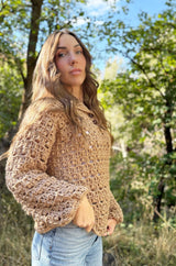 Side front view of woman wearing crochet pullover. It has long sleeves with a blousy cuff, button placket at top neck, and a lacy texture. The sweater is a warm tan beige.  thumbnail