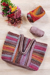 Tote bag with handles laid on top of bag, with yarn balls and flowers in the upper frame. The bag is made in shades of pink, purple, warm yellow, and steely grey blue. The middle panel is striped vertically and the side panels are striped horizontally.  thumbnail