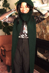 Woman stands in front of a stone fireplace and armchair, wearing a floral print shirt and grey pants. She has a crochet hood with a short cowl neck and long scarf extensions.  thumbnail