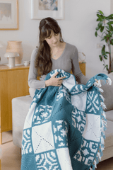 Woman sitting in a pale living room, on the arm of a couch. She has a crocheted afghan on her lap, made of multiple square patterns. It has fringe along the edge.  thumbnail