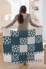 Woman standing in a pale room holds a folded crochet blanket. The blanket is made in white and medium blue, with solid squares in those colors and patterned squares mixing those colors. It had mixed fringe on the shorter top and bottom edges.  thumbnail